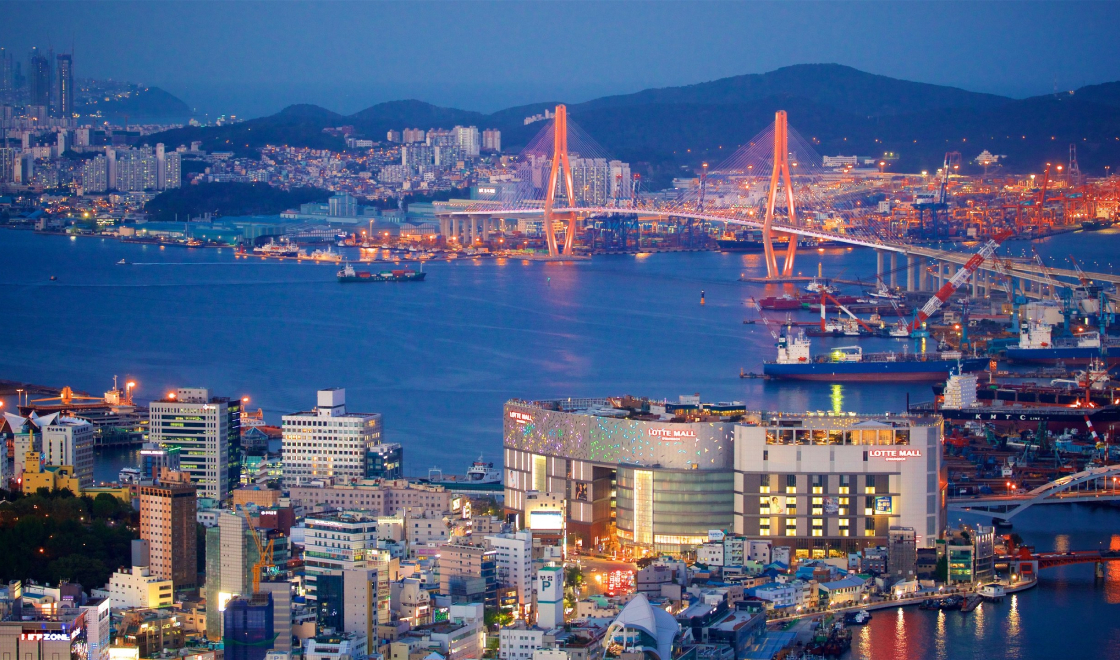 Vista panorâmica noturna de Busan, Coreia do Sul, com a ponte Gwangandaegyo iluminada ao fundo, refletindo suas luzes sobre a água. Em primeiro plano, está o Lotte Mall e edifícios ao redor, com diversas luzes destacando a vida urbana da cidade.