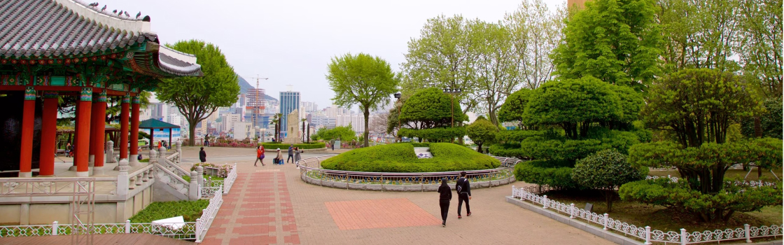 Vista do Parque Yongdusan em Busan, Coreia do Sul, com um pavilhão tradicional coreano à esquerda e uma paisagem de árvores podadas e caminhos pavimentados ao centro, emoldurados por vegetação exuberante e edifícios modernos ao fundo.