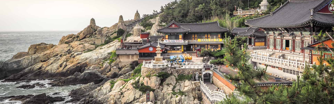Templo Haedong Yonggungsa em Busan, Coreia do Sul, localizado à beira-mar, com sua arquitetura tradicional coreana cercada por penhascos rochosos e vegetação. A cena destaca o templo contrastando com o oceano ao fundo, criando uma atmosfera de tranquilidade e beleza natural.